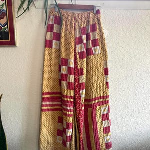 May include: Yellow patchwork pants with red and white accents. The pants feature a mix of patterns, including polka dots and checkered squares. The fabric appears to be quilted, with visible stitching throughout. The pants are hanging on a wooden hanger.