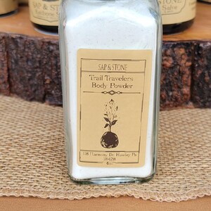 May include: A clear glass bottle with a brown wooden lid containing white body powder. The label reads SAP & STONE Trail Travelers Body Powder, 118 Harmony Dr. Hawley Pa 18428, 4oz, with a botanical illustration of a plant growing from a bulb.