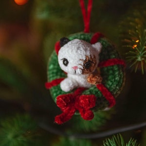 May include: A white crocheted cat ornament with brown eyes and a red bow sits inside a green crocheted wreath. The ornament is hanging on a Christmas tree.