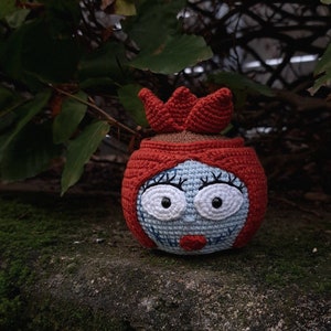 May include: A handmade crochet doll with a round head, blue face, and red hair. The doll has large white eyes, black eyelashes, and a red stitched mouth. The doll is sitting on a mossy stone.