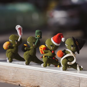 以下が含まれることがあります： 異なる休日の帽子をかぶった、緑色の毛糸で編まれた5体の恐竜の置物。恐竜たちは、ウサギ、クローバー、プレゼント、キャンディーケーン、魔女の帽子など、さまざまな休日にちなんだアイテムを持っています。