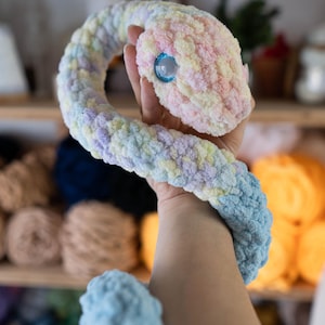 May include: A crocheted snake toy with a pastel color scheme, including pink, yellow, blue, and purple. The snake has a single blue eye and is curled in a loop.