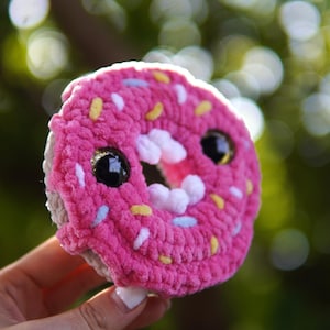 May include: A pink crocheted donut with white and yellow sprinkles and googly eyes. The donut is being held by a hand.