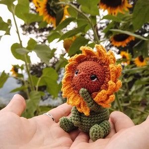 Puede incluir: Un juguete de peluche de girasol tejido a crochet con una cara marrón y ojos grandes, sostenido en las manos de una persona. El juguete de peluche está rodeado de girasoles reales.