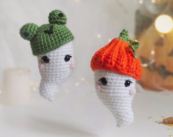 May include: Two white crocheted ghost ornaments with smiling faces. One ghost is wearing a green frog hat and the other is wearing an orange pumpkin hat.