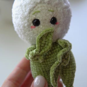 May include: A handmade plush toy with a white, fluffy head and a green body with leaf-like appendages. The toy has black eyes, pink cheeks, and a sweet expression. The plush is made of soft, textured material.