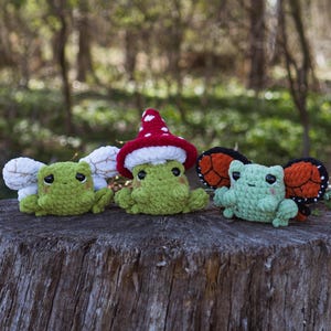 May include: Three handmade crochet frog plushies in shades of green, sitting on a tree stump. One frog wears a red mushroom cap with white polka dots. Another has white wings, and the third has orange and black butterfly wings.