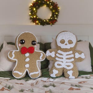 May include: Two festive plush pillows: a gingerbread man with a red bow tie and a skeleton. The gingerbread man is tan with white icing details. The skeleton pillow is white with a tan body. A Christmas wreath hangs above the pillows.