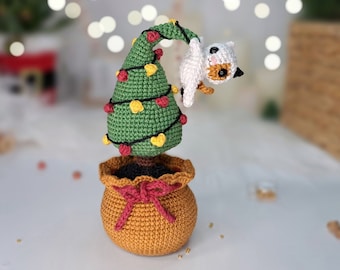 May include: A crocheted Christmas tree with red and yellow lights, a white cat with orange ears hanging from the top, and a brown pot with a red bow.