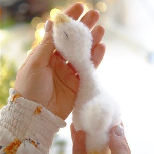 May include: A handmade, plush white goose with a yellow beak and feet. The soft toy is held gently in two hands, showcasing its fluffy texture. The goose is a charming, handcrafted item, perfect for a nursery or as a decorative piece.