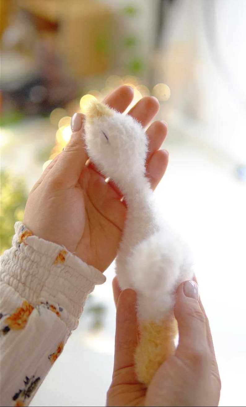 May include: A handmade, plush white goose with a yellow beak and feet. The soft toy is held gently in two hands, showcasing its fluffy texture. The goose is a charming, handcrafted item, perfect for a nursery or as a decorative piece.
