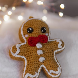 May include: A crocheted gingerbread man ornament with a red bow tie and white icing details. The ornament has black eyes and rosy cheeks. The background is a soft, white surface with blurred string lights.