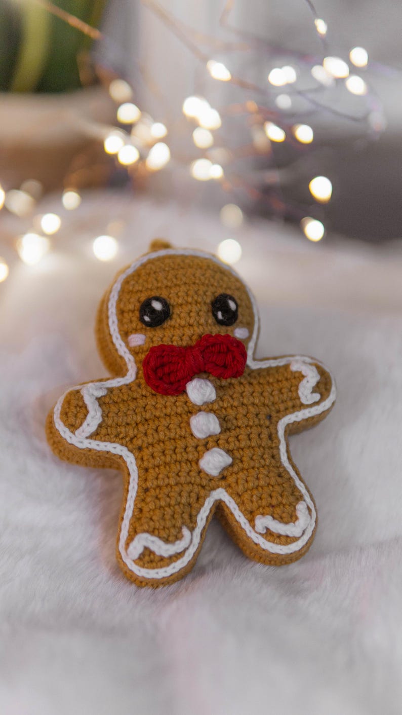 May include: A crocheted gingerbread man ornament with a red bow tie and white icing details. The ornament has black eyes and rosy cheeks. The background is a soft, white surface with blurred string lights.