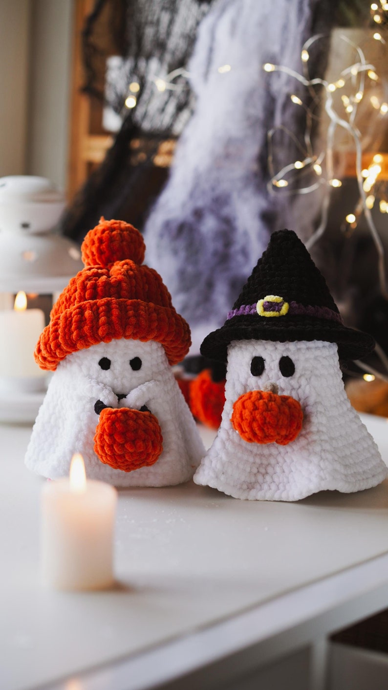 K&ouml;nnte beinhalten: Zwei geh&auml;kelte Geister-Ornamente, eines mit einem schwarzen Hexenhut und das andere mit einer leuchtend orangefarbenen Strickm&uuml;tze. Beide Geister halten orangefarbene Wollk&uuml;rbisse in ihren H&auml;nden.