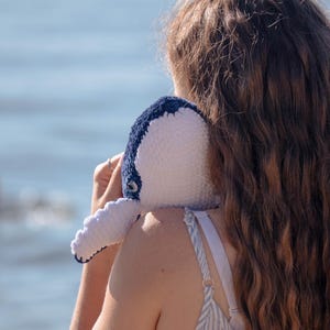 K&ouml;nnte beinhalten: Ein handgefertigtes Wal-Pl&uuml;schtier aus H&auml;kelarbeit in Wei&szlig; und Marineblau, mit detailliertem Auge und Flosse. Das Pl&uuml;schtier wird auf der Schulter einer Person gehalten, mit langen braunen Haaren und einem blau-wei&szlig; gestreiften Kleid.