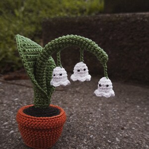 May include: A crocheted lily of the valley plant in a terracotta pot. The plant has white flowers with black eyes and green leaves.
