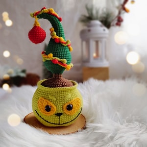 May include: A crocheted Christmas tree with red and yellow ornaments in a green and yellow pot with a smiling face.