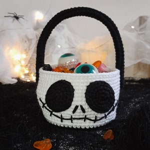 K&ouml;nnte beinhalten: Wei&szlig;er geh&auml;kelter Halloween-Trick-or-Treat-Korb in Form eines Sch&auml;dels mit schwarzen Augen und einem gestickten L&auml;cheln. Der Korb ist gef&uuml;llt mit S&uuml;&szlig;igkeiten und Spielzeugaugen.