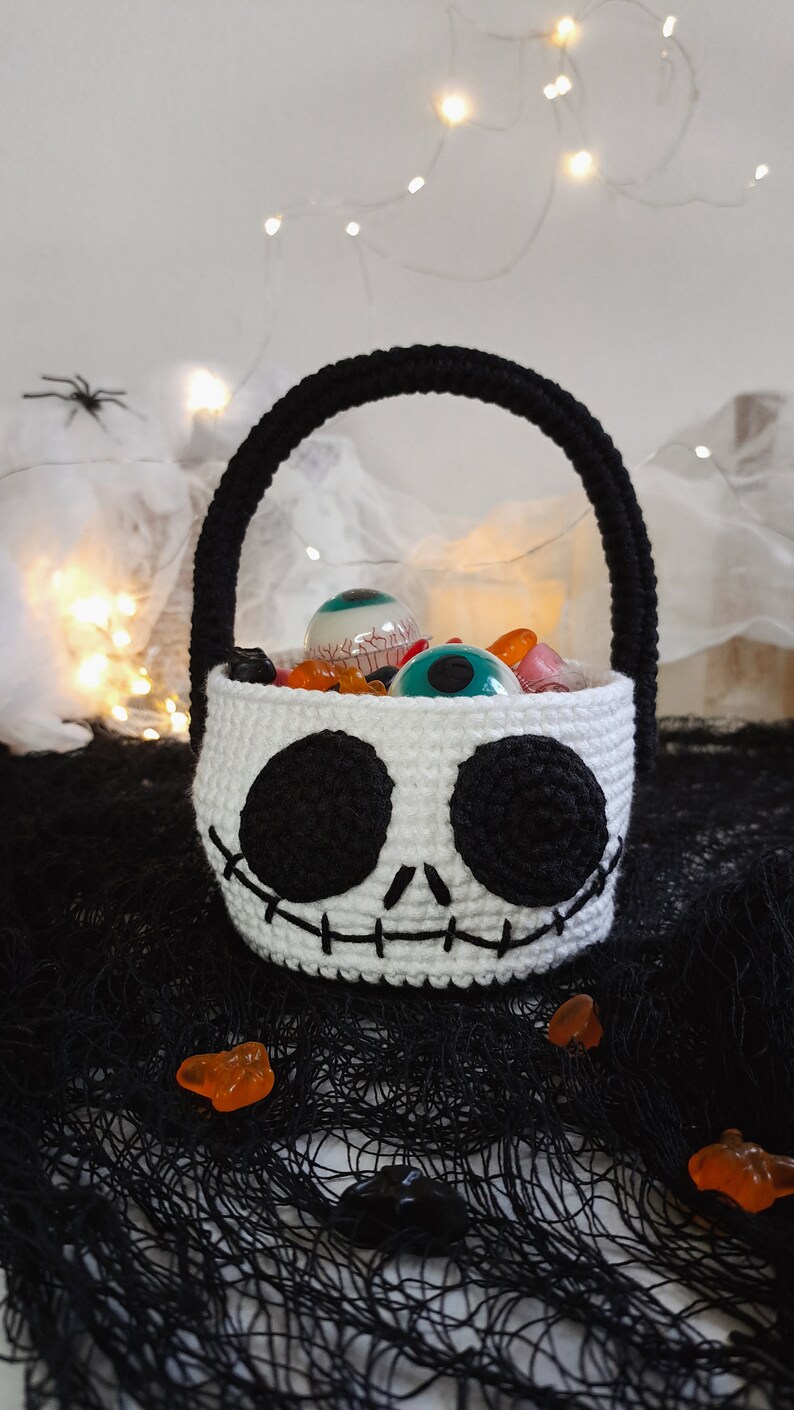 K&ouml;nnte beinhalten: Wei&szlig;er geh&auml;kelter Halloween-Trick-or-Treat-Korb in Form eines Sch&auml;dels mit schwarzen Augen und einem gestickten L&auml;cheln. Der Korb ist gef&uuml;llt mit S&uuml;&szlig;igkeiten und Spielzeugaugen.