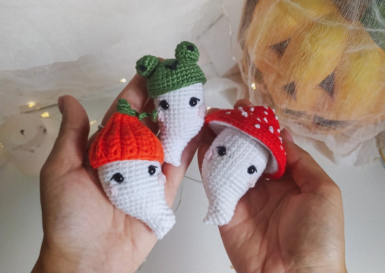 May include: Three crocheted ghost ornaments with different hats. One ghost wears a green frog hat, another wears an orange pumpkin hat, and the third wears a red mushroom hat.
