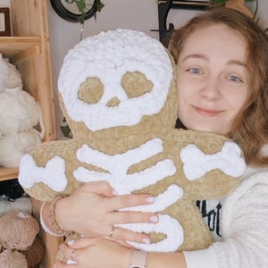 May include: A plush, tan-colored skeleton-shaped pillow with white bone detailing. The pillow is soft and cuddly, perfect for Halloween or spooky decor. The skull and bones are clearly defined, creating a fun, whimsical design.