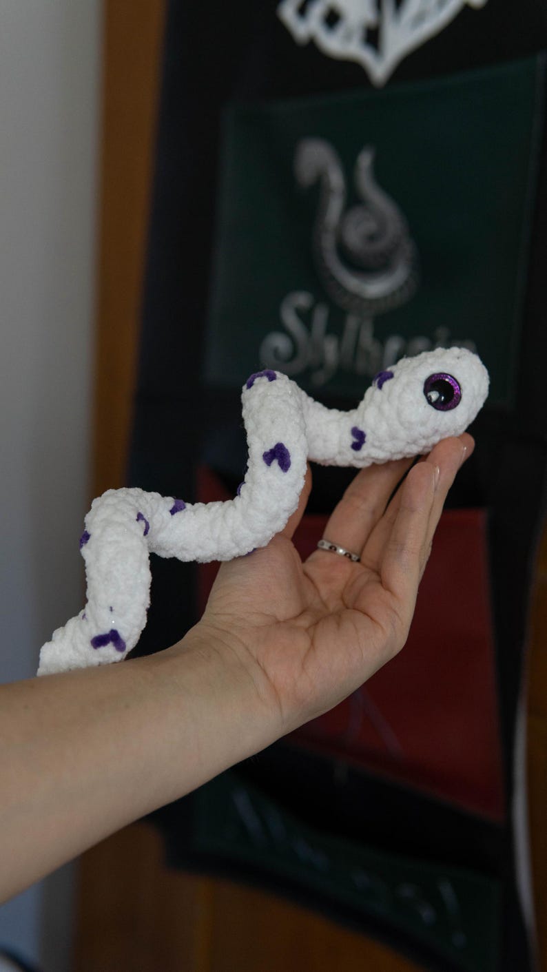 K&ouml;nnte beinhalten: Eine wei&szlig;e geh&auml;kelte Schlange mit violetten Akzenten und glitzernden violetten Augen. Die Schlange ist in der Hand einer Person zusammengerollt.