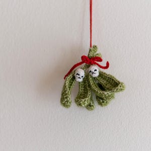 Pu&ograve; includere: Un ornamento di vischio in crochet verde con due bacche in crochet bianco e un nastro rosso.