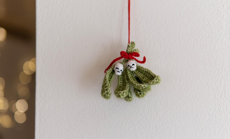 Pu&ograve; includere: Un ornamento di vischio in crochet verde con due bacche in crochet bianco e un nastro rosso.
