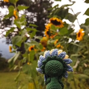 K&ouml;nnte beinhalten: Ein geh&auml;keltes Sonnenblumen-Amigurumi-Spielzeug mit gr&uuml;nem K&ouml;rper, blauen und gelben Bl&uuml;tenbl&auml;ttern und einer braunen Mitte. Das Spielzeug wird in einer Hand vor einem Hintergrund aus echten Sonnenblumen gehalten.