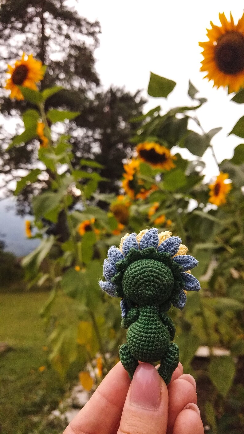 K&ouml;nnte beinhalten: Ein geh&auml;keltes Sonnenblumen-Amigurumi-Spielzeug mit gr&uuml;nem K&ouml;rper, blauen und gelben Bl&uuml;tenbl&auml;ttern und einer braunen Mitte. Das Spielzeug wird in einer Hand vor einem Hintergrund aus echten Sonnenblumen gehalten.