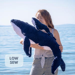 Puede incluir: Dos ballenas de peluche, una grande y otra pequeña, en tonos azul marino y blanco. La ballena más grande está sujeta por una persona. Las ballenas están hechas de un material suave y texturizado. La imagen incluye el texto "LOW SEW".
