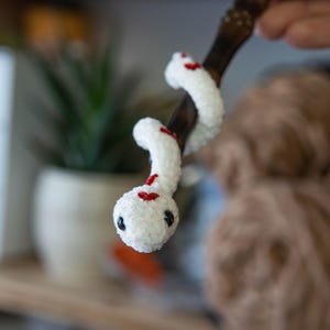 K&ouml;nnte beinhalten: Eine wei&szlig;e geh&auml;kelte Schlange mit roten Akzenten, die um einen Holzstab gewickelt ist. Die Schlange hat schwarze Augen und eine strukturierte Oberfl&auml;che.