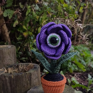 Puede incluir: Una flor de color morado tejida a crochet con un ojo verde en el centro, en maceta de color marr&oacute;n. La flor est&aacute; sentada sobre un tronco de madera.