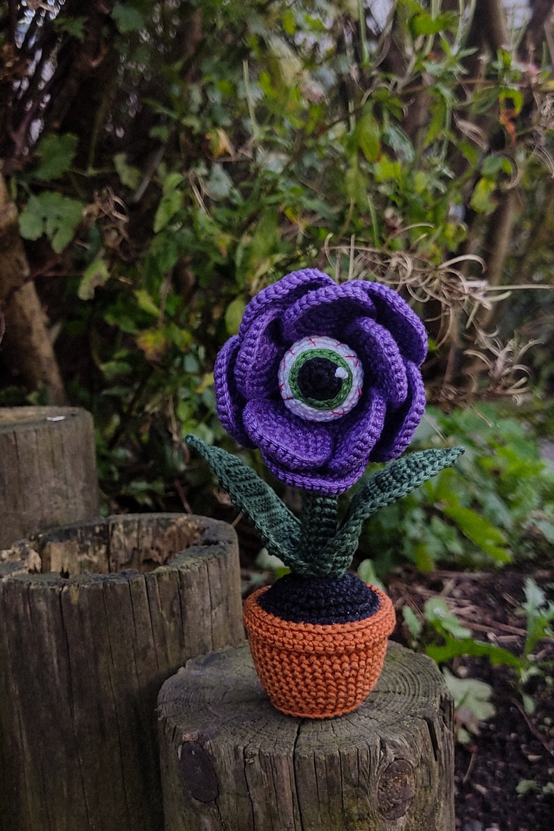 Puede incluir: Una flor de color morado tejida a crochet con un ojo verde en el centro, en maceta de color marr&oacute;n. La flor est&aacute; sentada sobre un tronco de madera.