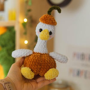 Könnte beinhalten: Ein handgemachtes Plüschtier in Form einer Gans. Sie hat einen weißen Kopf und Flügel, einen gelben Schnabel und Füße sowie einen orangefarbenen Körper und Hut. Die Gans wird in einer Hand gehalten. Es ist ein weiches, kuscheliges Spielzeug.