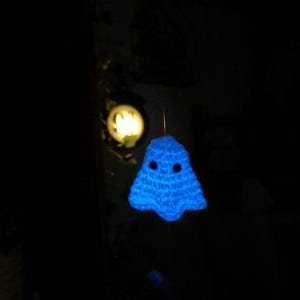 Puede incluir: Una decoraci&oacute;n de fantasma azul brillante tejida a ganchillo con ojos de bot&oacute;n negros cuelga frente a un fondo oscuro. Un objeto redondo e iluminado es visible en el fondo. El fantasma tiene un dise&ntilde;o simple y amigable.