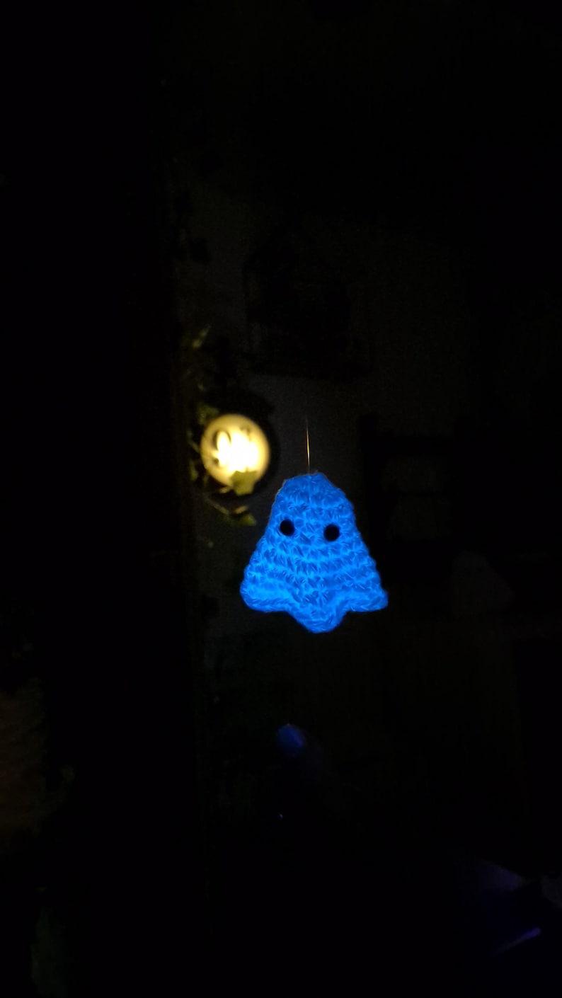 Puede incluir: Una decoraci&oacute;n de fantasma azul brillante tejida a ganchillo con ojos de bot&oacute;n negros cuelga frente a un fondo oscuro. Un objeto redondo e iluminado es visible en el fondo. El fantasma tiene un dise&ntilde;o simple y amigable.