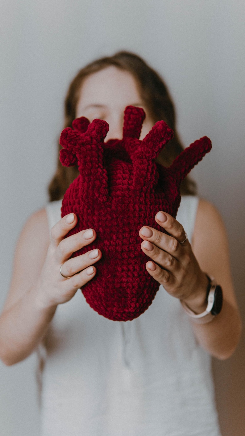 May include: A person holding a large, red crocheted heart in front of their face.