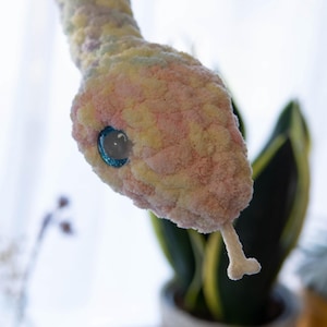 K&ouml;nnte beinhalten: Ein geh&auml;keltes Schlangenspielzeug in Pastellrosa, Gelb und Blau. Die Schlange hat ein gro&szlig;es, blaues Auge und eine wei&szlig;e Zunge.