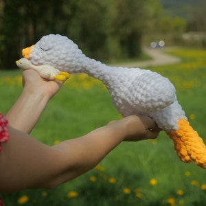 May include: A large white crochet goose with yellow feet and beak, and a smaller matching goose. The larger goose is lying down, appearing to be sleeping. The background is a green field with yellow flowers.