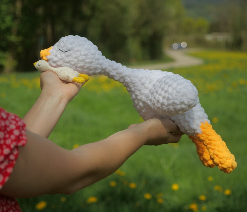 May include: A large white crochet goose with yellow feet and beak, and a smaller matching goose. The larger goose is lying down, appearing to be sleeping. The background is a green field with yellow flowers.