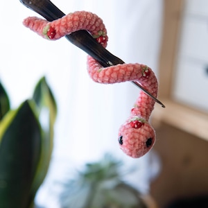 K&ouml;nnte beinhalten: Eine rosa geh&auml;kelte Schlange mit roten und gr&uuml;nen Akzenten ist um eine Stricknadel aus Holz gewickelt.