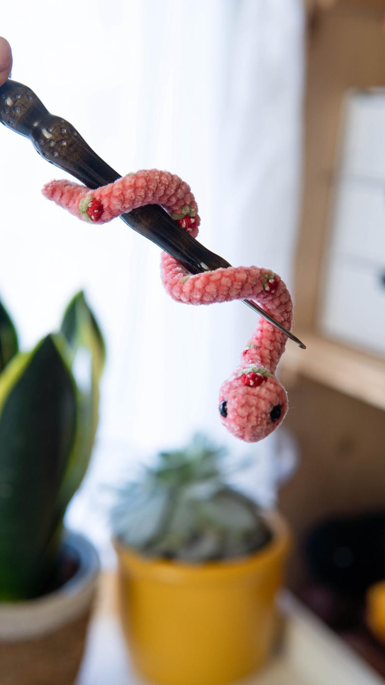 K&ouml;nnte beinhalten: Eine rosa geh&auml;kelte Schlange mit roten und gr&uuml;nen Akzenten ist um eine Stricknadel aus Holz gewickelt.