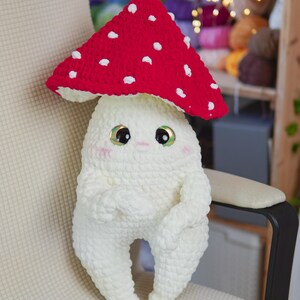 以下が含まれることがあります： 赤い帽子に白い水玉模様の、手作りのキノコのおもちゃ。キノコは白い体、腕、脚、そして大きく表情豊かな目をしています。おもちゃは椅子に座っています。