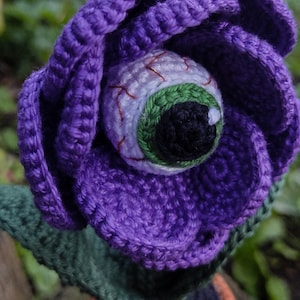 Puede incluir: Una flor de color morado tejida a crochet con un ojo verde y blanco en el centro. La flor est&aacute; sentada sobre un tallo verde tejido a crochet y una maceta marr&oacute;n.