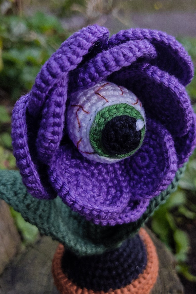 Puede incluir: Una flor de color morado tejida a crochet con un ojo verde y blanco en el centro. La flor est&aacute; sentada sobre un tallo verde tejido a crochet y una maceta marr&oacute;n.