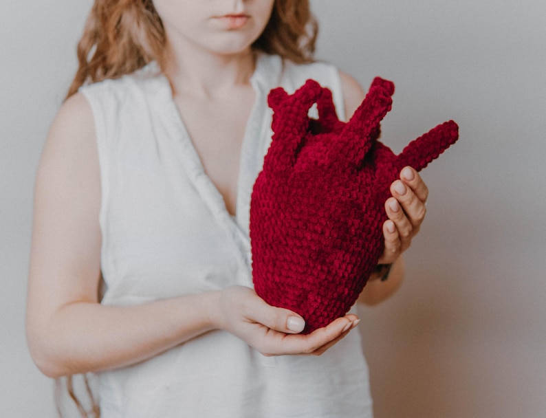 May include: A person holding a large, red crocheted heart. The heart is made of soft, fuzzy yarn and has a realistic shape.