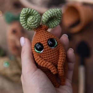 May include: A crocheted mandrake doll with a brown body and green leaves. The doll has large black eyes with green highlights. The doll is held in a hand.