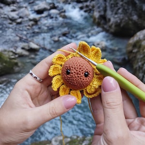 K&ouml;nnte beinhalten: Eine geh&auml;kelte Sonnenblume mit einem braunen Gesicht und schwarzen Augen wird mit einer H&auml;kelnadel bearbeitet. Die Sonnenblume ist gelb und hat einen gr&uuml;nen Stiel.