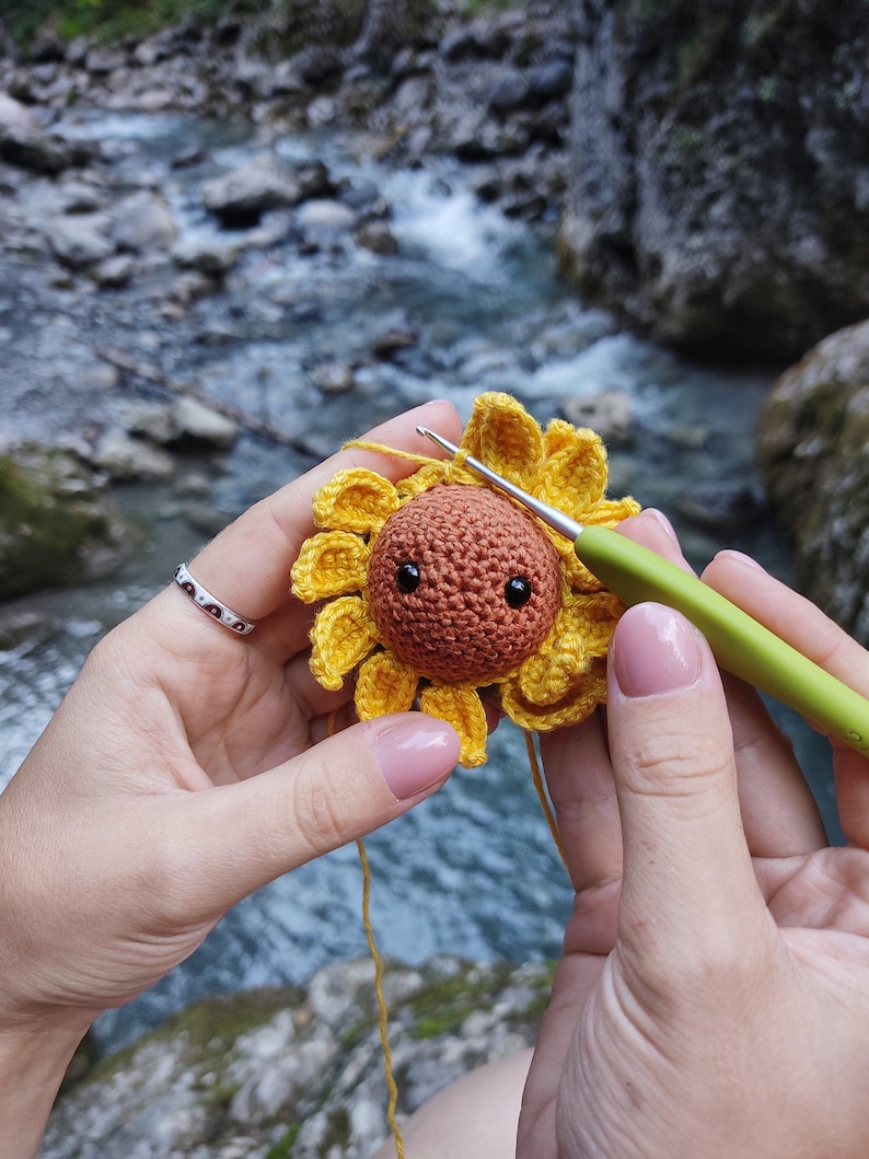 K&ouml;nnte beinhalten: Eine geh&auml;kelte Sonnenblume mit einem braunen Gesicht und schwarzen Augen wird mit einer H&auml;kelnadel bearbeitet. Die Sonnenblume ist gelb und hat einen gr&uuml;nen Stiel.
